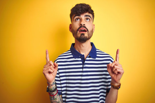 Young man with tattoo wearing striped polo standing over isolated yellow background amazed and surprised looking up and pointing with fingers and raised arms.