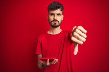 Young man with tattoo using tablet standing over isolated red background with angry face, negative...