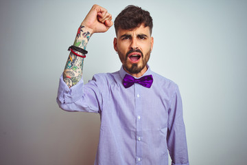 Young man with tattoo wearing purple shirt and bow tie over isolated white background angry and mad raising fist frustrated and furious while shouting with anger. Rage and aggressive concept.