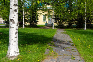 Stone-paved path to small yellow house among birches and firs