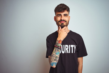 Young safeguard man with tattoo wering security uniform over isolated white background looking confident at the camera with smile with crossed arms and hand raised on chin. Thinking positive.