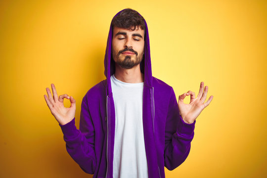 Young man with tattoo wearing purple sweatshirt with hood over isolated yellow background relax and smiling with eyes closed doing meditation gesture with fingers. Yoga concept.