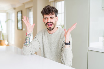 Young man wearing casual sweater sitting on white table celebrating mad and crazy for success with...