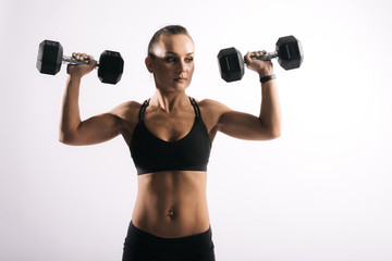 Portrait of muscular sexy young woman in black sportswear lifting dumbbells at shoulder level on isolated white background.