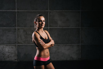 Obraz premium Fitness muscular woman with perfect muscular body in black sportswear is standing with arms folded, on dark isolated background