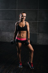 Young muscular woman bodybuilder with perfect athletic body in sports black bra is posing with two dumbbells on black isolated background. Sporty muscular girl looking at the camera