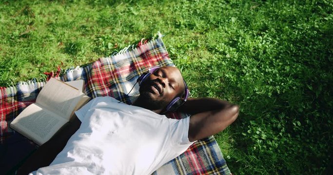 Happy african-american male student lying in park, listening to music in earphones and smiling, rela[