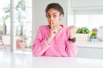 Fototapeta na wymiar Beautiful african american woman with afro hair wearing casual pink sweater asking to be quiet with finger on lips pointing with hand to the side. Silence and secret concept.
