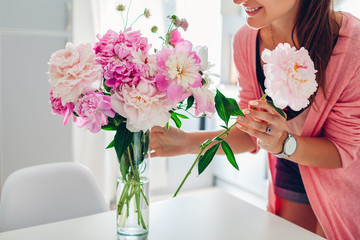 Woman puts peonies flowers in vase. Housewife taking care of coziness and decor on kitchen. Composing bouquet. © maryviolet