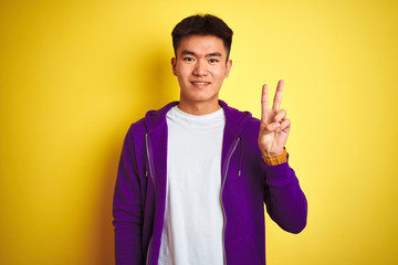 Young asian chinese man wearing purple sweatshirt standing over isolated yellow background showing and pointing up with fingers number two while smiling confident and happy.