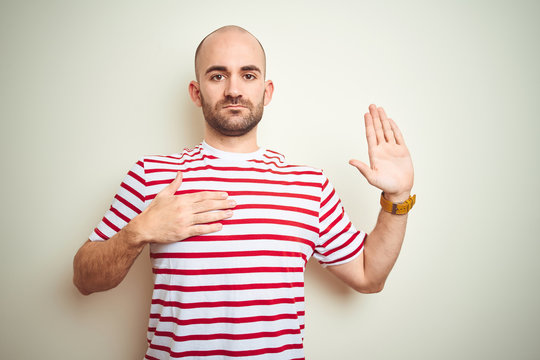 Young bald man with beard wearing casual striped red t-shirt over white isolated background Swearing with hand on chest and open palm, making a loyalty promise oath