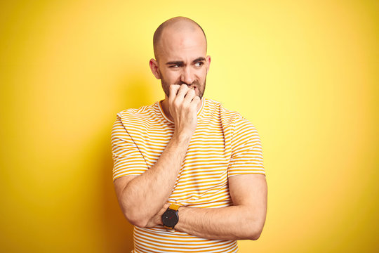 Young bald man with beard wearing casual striped t-shirt over yellow isolated background looking stressed and nervous with hands on mouth biting nails. Anxiety problem.