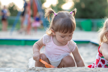 Naklejka premium Cute toddler girl playing in sand on outdoor playground. Beautiful baby in red trousers having fun on sunny warm summer day. Child with colorful sand toys. Healthy active baby outdoors plays games