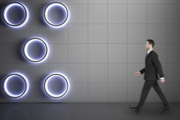 Businessman walking on concrete floor along blank grey wall with circle lights, mock up.