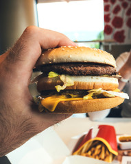 Male hand holding delicious double hamburger admiring the high calorie meat and bun with salad and cheese in between