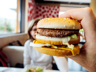 Male hand holding delicious double hamburger admiring the high calorie meat and bun with salad and cheese in between with woman defocused silhouette in background