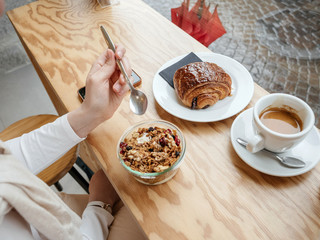 Side overhead view of single woman eating delicious yogurt with muesli drinking coffee with croissant and cake early in the morning in French cafe in Chambery