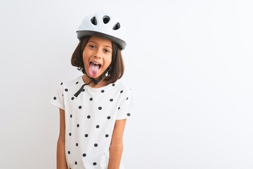 Beautiful child girl wearing security bike helmet standing over isolated white background sticking tongue out happy with funny expression. Emotion concept.