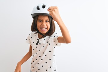 Beautiful child girl wearing security bike helmet standing over isolated white background angry and mad raising fist frustrated and furious while shouting with anger. Rage and aggressive concept.