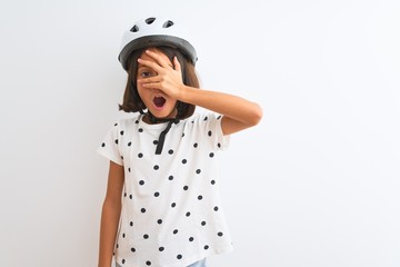 Beautiful child girl wearing security bike helmet standing over isolated white background peeking in shock covering face and eyes with hand, looking through fingers with embarrassed expression.