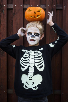 Happy young blond hair boy with skeleton costume holding jack o lantern. Halloween. Trick or treat. Outdoors portrait over wooden background