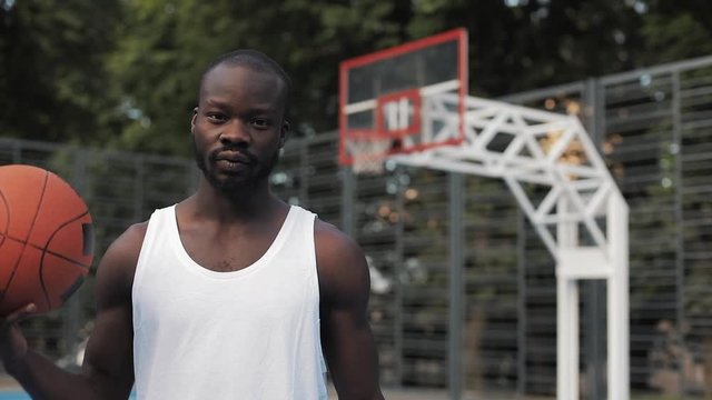 Close Up Portrait of Young Serious Muscly Afro - American Guy in White Singlet Playing with ball, Standing at Street Basketbal Court and Looking to Camera. Healthy Lifestyle and Sport Concept.