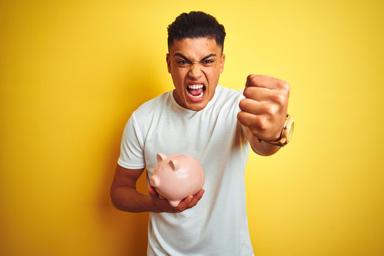 Young brazilian man holding piggy bank standing over isolated yellow background annoyed and frustrated shouting with anger, crazy and yelling with raised hand, anger concept