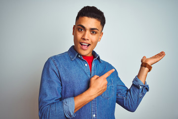 Young brazilian man wearing denim shirt standing over isolated white background amazed and smiling to the camera while presenting with hand and pointing with finger.