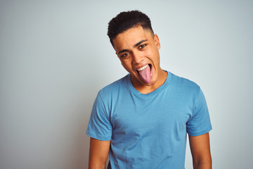 Young brazilian man wearing blue t-shirt standing over isolated white background sticking tongue out happy with funny expression. Emotion concept.