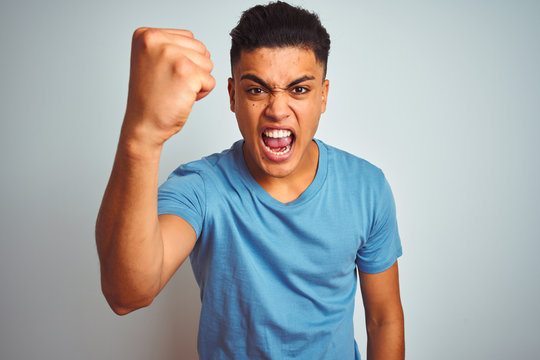 Young brazilian man wearing blue t-shirt standing over isolated white background angry and mad raising fist frustrated and furious while shouting with anger. Rage and aggressive concept.