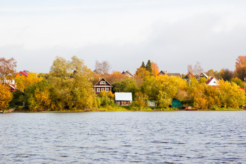 Beautiful autumn landscape with wooden private houses and agricultural buildings on riverbank, colorful autumn trees and blue sky. Outdoor recreation and countryside life concept. Autumn scene.