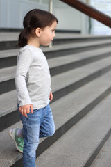 Obraz premium little cute white girl brunette with pigtails in jeans standing on the stairs
