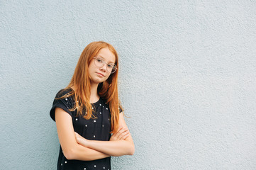 Outdoor portrait of sweet little girl with red hair, wearing eyeglasses, arms crossed