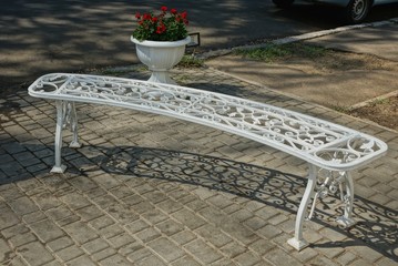 a white metal bench with a forged pattern stands on a gray sidewalk