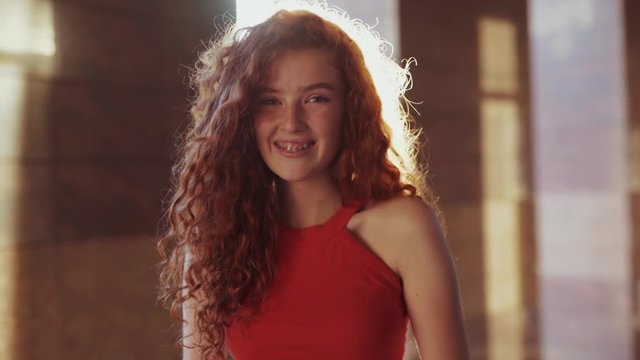 Extremely beautiful red headed curly young Caucasian student girl in trendy red dress looks straight to camera and laughs happily in bright sunlight outdoors. Female portrait, close up view, facial