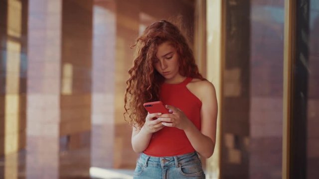 Close up view of young sweet gorgeous student girl in trendy red shirt and jeans standing outdoors by the college, office, school and actively chatting via online messaging app. Modern technology
