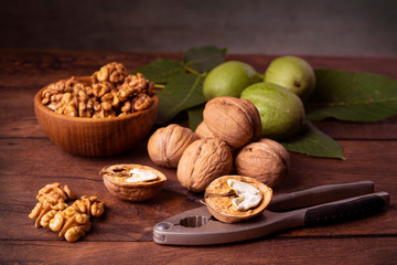 Walnuts chopped in a bowl, whole and in a green peel and a nut cracker on a dark old wooden background.