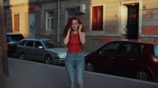 Beautiful young curly ginger woman walking down the street and listening to music from smartphone in earphones, dancing passionately. Beautiful teenager girl walking and dancing. Female portrait