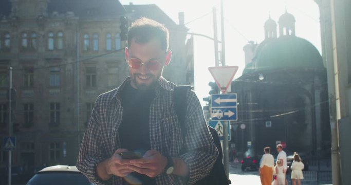 Trendy young man in sunglasses reading dunny news on smartphone walking in tourist street of gorgeous city on sunny spring day.