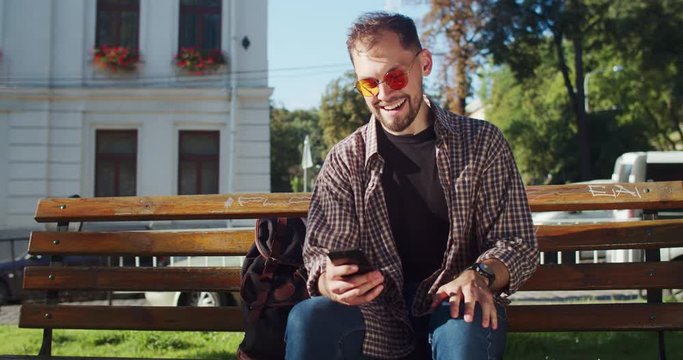 Happy young man learning exciting news using smartphone clenching fist like enjoying success or victory sitting on bech in the downtown.