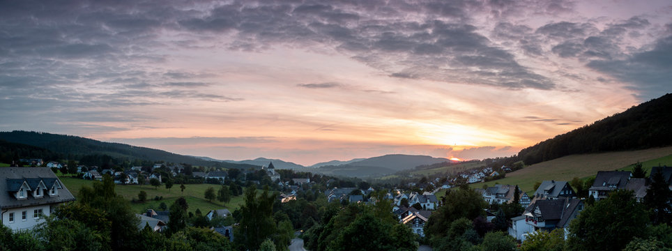 Wide panoramic view over the mountainous village of Grafschaft in the winter sports region of Sauerland, Germany, during sunset with a partly colourful orange lit cloudy sky over the valley