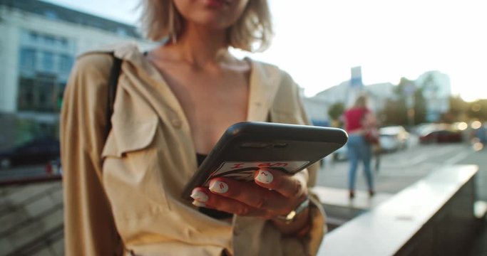 Sociable young woman with blond ehair holdin smartphone browsing in social media checking news staying outdoor in moden big city.