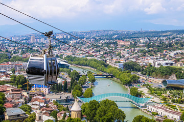View of cable car above Tbilisi Georgia with view of Mtkvari - Kura River and Peace Bridge and city with mountains in the distance - obrazy, fototapety, plakaty