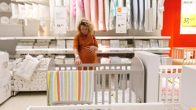 Young Pregnant Woman Choosing Cot Or Baby Bed In Furniture Store.