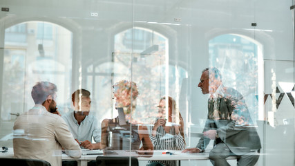 Fototapeta na wymiar Office life. Group of business people discussing something while sitting at the office table behind the glass wall in the modern office
