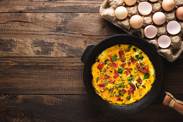 Top view omelette with tomatoes and greens, pumpkin seeds in cast iron rustic frying pan on dark wooden background with eggs in cardboard container with copy space