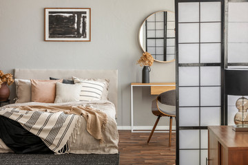 Elegant round mirror in wooden frame above fancy console table with flowers in vase in trendy bedroom interior with beige vase