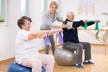 Zelfklevend Fotobehang Elderly man and woman exercising on gymnastic balls during physiotherapy session at hospital © Photographee.eu