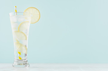 Healthy fresh tonic with lemon, ice cubes, soda, yellow striped straw in misted glass on white wood table, pastel green color background.