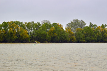 Fototapeta na wymiar Athlete paddling SUP (Stand up paddle board) at Danube river at cold weather against overcast sky. Concept of water tourism, water sport, healthy lifestyle and recreation